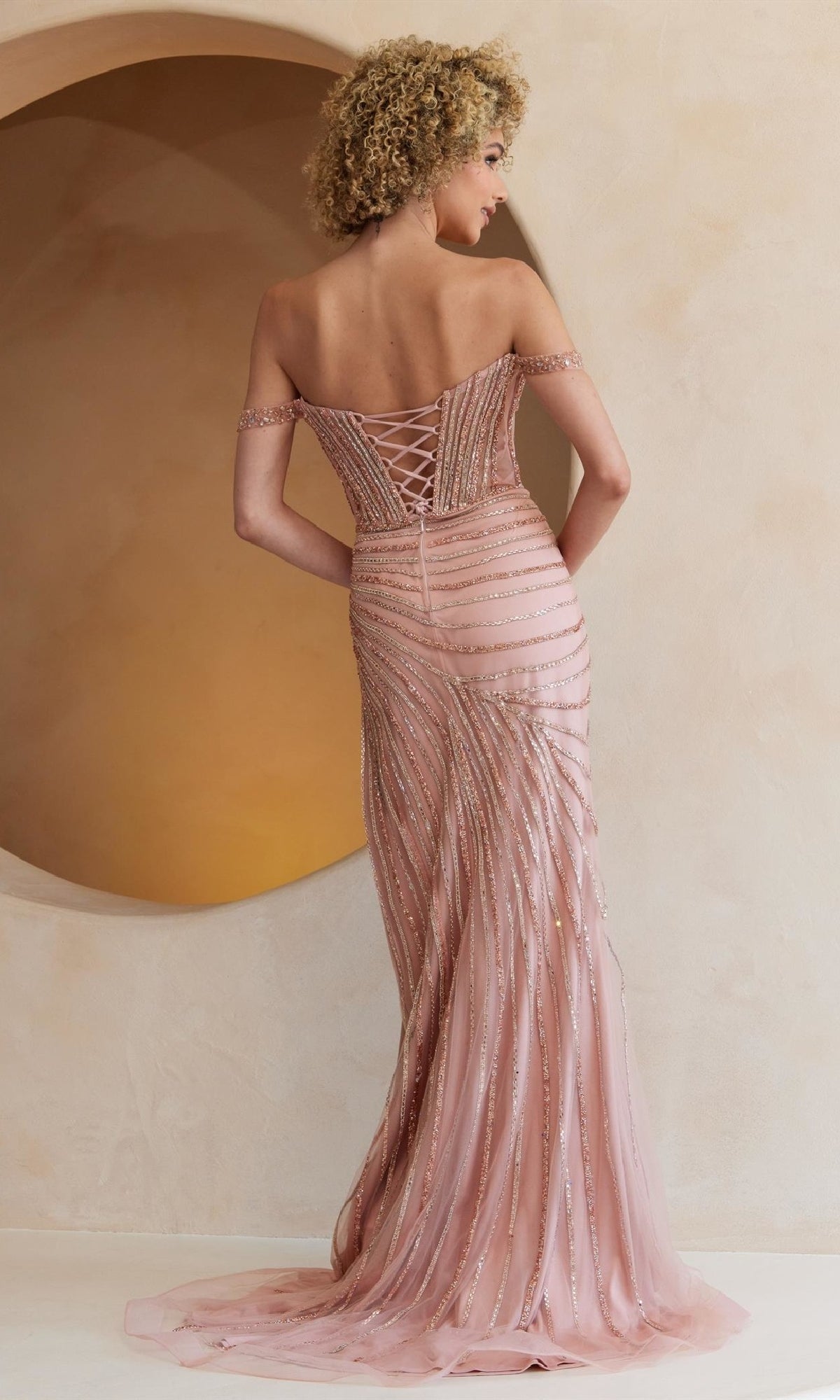 A woman with curly hair models the Beaded Long Gold Prom Dress (Amelia Couture TM1029), featuring beaded details and corset-style lacing on the back, standing in front of a beige wall.